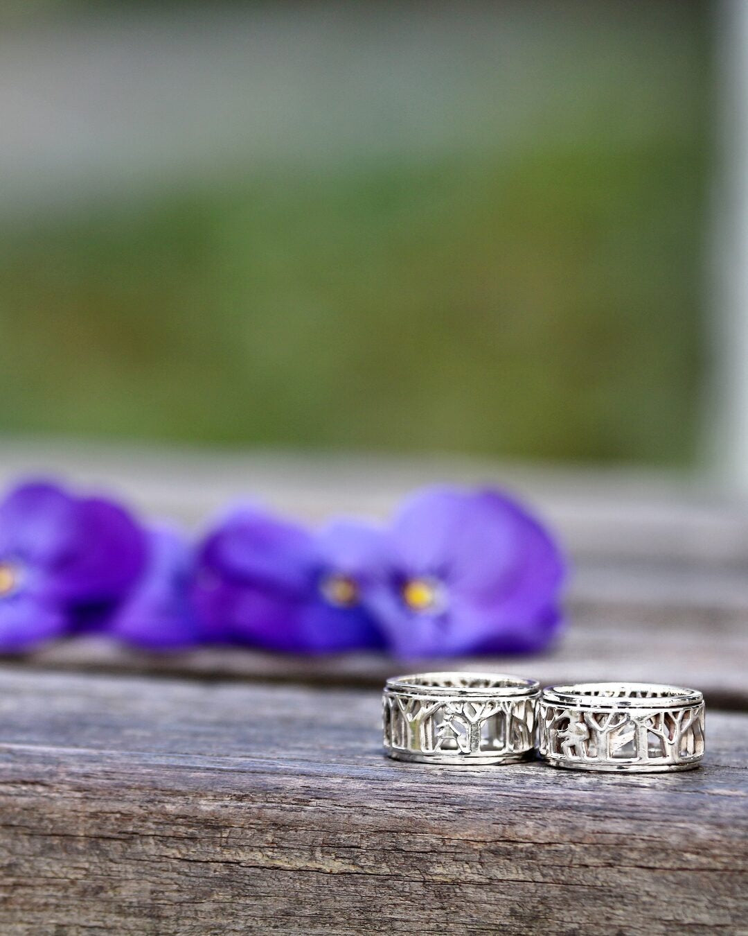 Two silver spinner rings with a forest cutout on a wooden table beside violets